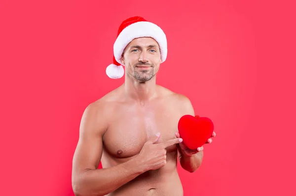 xmas heart and man with torso pointing finger. shirtless man in xmas santa claus hat with heart gift. xmas man isolated on red background. man hold heart at xmas in studio.