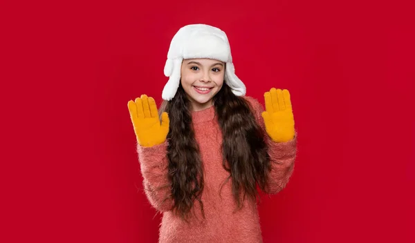 冬季针织物 戴耳瓣帽的少女模特儿举手 戴着冬季帽子的少女被红色背景隔离了 在演播室里 少女穿着毛衣 头戴冬帽 少女冬季时装 — 图库照片