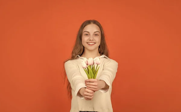 happy kid hold flowers for spring holiday. teen girl with bouquet on orange background. childhood happiness. floral present for womens day. march day. child with tulips.