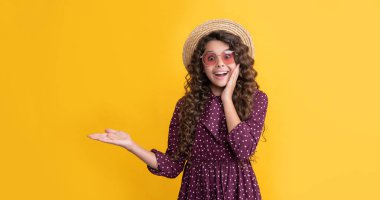 Hasır şapkalı ve güneş gözlüklü uzun kıvırcık saçlı çocuk ürününü sunuyor..