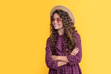 Sarı arka planda uzun sarı kıvırcık saçlı, güneş gözlüklü, saman şapkalı pozitif çocuk..