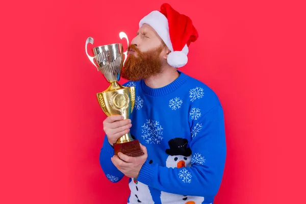 Bearded Santa man kissing champion trophy cup studio isolated on red. Celebrating achievement. Merry Christmas. Happy New Year. Holidays celebration.