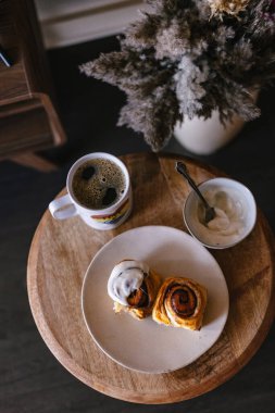 Beyaz vanilyalı kremalı tarçınlı çörek. Huysuz siyah yemek fotoğrafı. Kahve ve pişmiş hamur işi kahvaltı. Taze pişirme ikramı. Kahverengi tarçın şekeri. Yuvarlak girdaplar. Kahve masası. Brunch.
