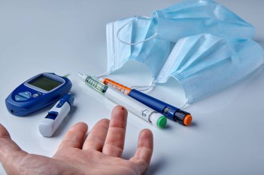 Male hand with a drop of blood on a finger, a glucometer, insulin syringe pens and medical masks on a white table. Diabetes and coronavirus clipart