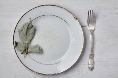 Beyaz ahşap bir masanın üzerinde kurumuş yapraklarla süslenmiş çatallı eski beyaz bir tabak. Yemek gösterisi için maket.