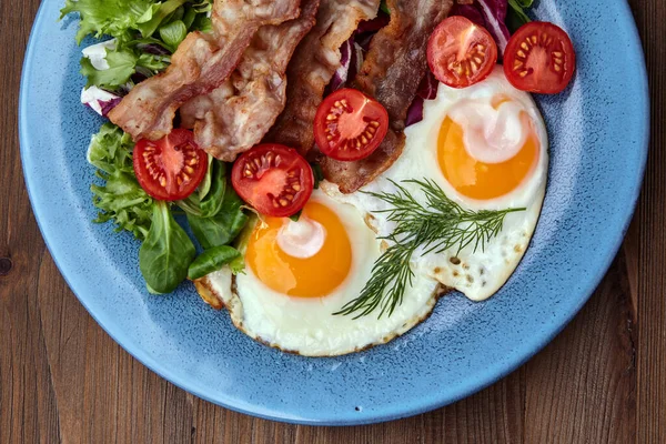 Fried eggs on a blue plate with bacon, lettuce and cherry tomatoes — Stock Photo, Image
