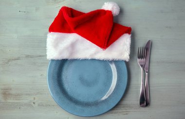 Mavi ahşap bir masa üzerinde Noel şapkası ve çatal bıçak takımı olan boş mavi tabak.