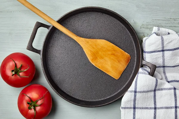 Boş yuvarlak dökme demir tava, ahşap spatula, çay havlusu ve domates.