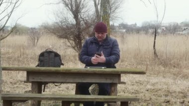 Doğada bir adam şehrin arka planında bir masada otururken telefonuna bakar.
