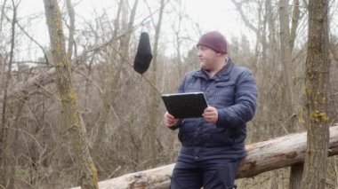  Doğada bir adam devrilmiş bir çam ağacında otururken tabletindeki fotoğraflara bakar. Koruyucu bir maske bir dala asılı..