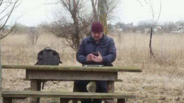 Doğada bir adam şehrin arka planına karşı masada otururken telefonundaki fotoğraflara bakar.