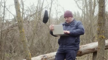 Ormanda serbest çalışan biri, devrilmiş bir çam ağacının üzerinde otururken sol eliyle bir dizüstü bilgisayarla yazı yazıyor. Koruyucu bir maske bir dala asılı..