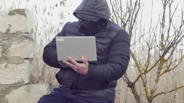Mężczyzna Kapturem Głowie Pisze Coś Laptopie Siedząc Zawalonej Ścianie — Wideo stockowe