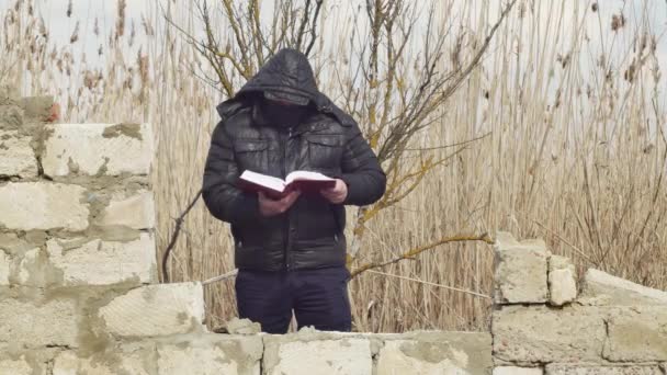 頭にフードを被った男が倒壊した壁の後ろに立っている厚い本を読む — ストック動画