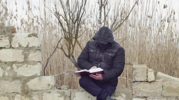 Человек Капюшоном Голове Читает Библию Сидя Разрушенной Стене — стоковое видео