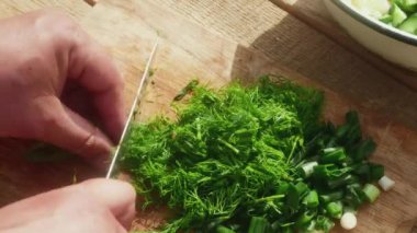 a hand with a knife cuts dill on a wooden table for salad, close-up