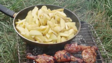 a hand with a fork mixes potatoes in a skillet at the camping barbecue grill, and checks for readiness of chicken drumsticks.