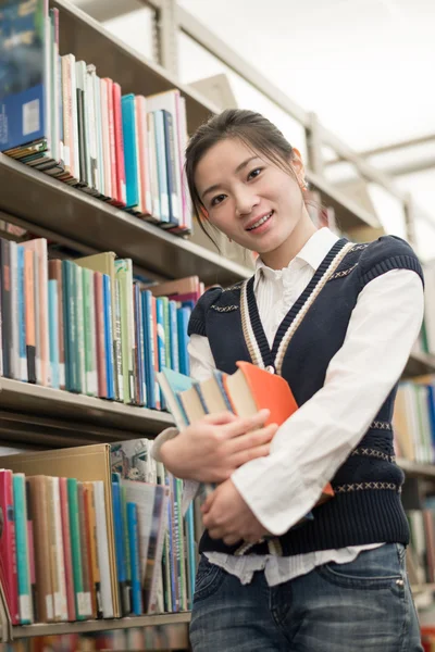 Student holding books next to bookshelf — Stock Photo, Image