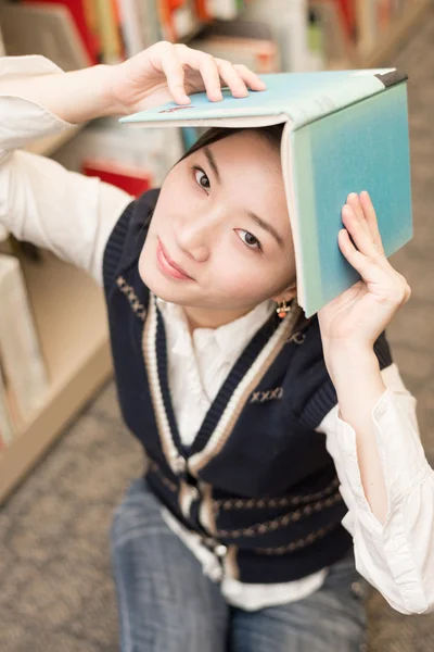 Chica sosteniendo un libro sobre la cabeza cerca de la estantería —  Fotos de Stock