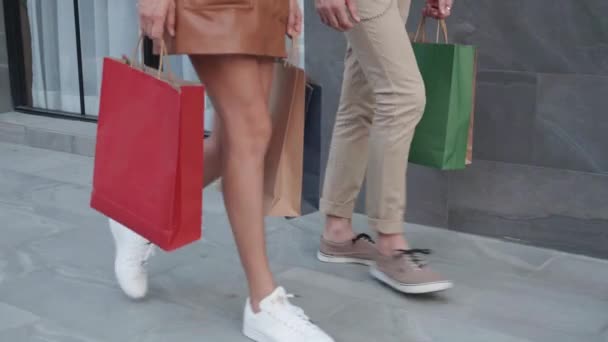 2人の若い男性と女性の閉鎖脚を歩いて通りで買い物袋を保持し 友人が一緒に購入するためのお店ですが カップルの関係は満足して消費主義です — ストック動画