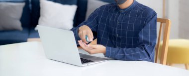 Genç Asyalı erkek evde alkol jeli ya da dezenfektanla çalışır. Coronavirüs ya da covid-19, yeni normal karantina ve sosyal mesafe, dizüstü bilgisayar kullanan iş adamı..