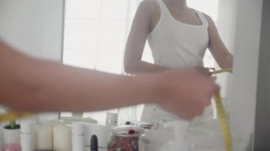 Güzel, genç Asyalı bir kadın ayakta duruyor ve ölçü alıyor. Kilo vermek için beliyle. Odada bir ayna var..