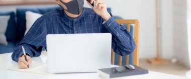 Yüz maskeli Asyalı genç bir iş adamı akıllı telefondan konuşuyor ve bilgisayarla covid-19 'u koruyor, iş adamı karantina işini evden yapıyor ve not defteri yazıyor, sosyal mesafe koyuyor ve evde kalıyor..