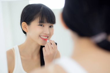 Güzel, genç Asyalı bir kadın dudaklarına ruj sürüyor, dudaklarına kırmızı ruj sürüyor, güzellik kızı makyaj malzemesi için aynaya bakıyor, zarif dudaklarıyla makyaj yapıyor, seksi bir kadın, cilt bakımı ve sağlık konseptiyle barışıyor..