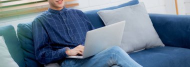 Genç Asyalı iş adamı evde dizüstü bilgisayarla gülüyor ve çalışıyor oturma odasındaki kanepede internete bağlanıyor, serbest çalışan adam kanepeye dizüstü bilgisayar kullanıyor, konforlu, yeni normal yaşam tarzı konsepti..