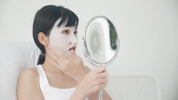 漂亮的年轻亚洲女人 戴着薄片面膜 在家里照镜子 漂亮的女孩用化妆品和化妆品 用皱纹护肤 护肤和保湿霜触摸脸 — 图库视频影像