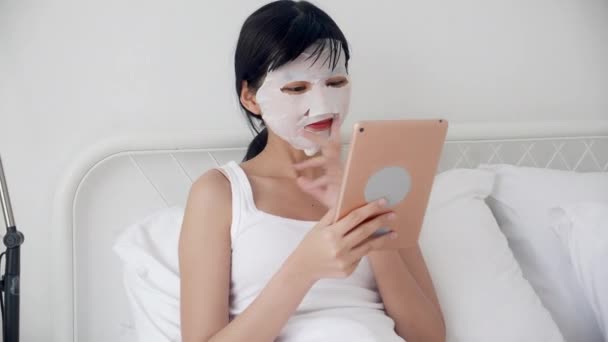漂亮的年轻亚洲女人 戴着薄片面罩 坐在床上看平板电脑 漂亮的女孩在家里用面罩护肤 降和健康 — 图库视频影像