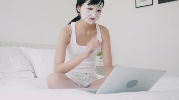 漂亮的年轻亚洲女人 戴着薄片面膜 坐在床上的笔记本电脑上工作 漂亮的女孩在家里用面膜护肤 降和健康 — 图库视频影像