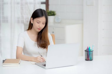 Genç Asyalı kadın dizüstü bilgisayarı evde çalışırken ve kullanırken planlamak için deftere yazı yazıyor, kız notları finans, kadın çalışmaları ve öğrenimi, iş ve iletişim konsepti hakkında.