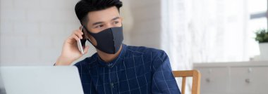 Yüz maskeli Asyalı genç bir iş adamı akıllı telefondan konuşuyor ve bilgisayar başında Coperid-19 'u korumak için çalışıyor. İş adamı karantina işini evden defter ve sosyal mesafeyle yapıyor ve evde kalıyor..