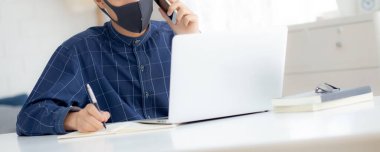 Yüz maskeli Asyalı genç bir iş adamı akıllı telefondan konuşuyor ve bilgisayarla covid-19 'u koruyor, iş adamı karantina işini evden yapıyor ve not defteri yazıyor, sosyal mesafe koyuyor ve evde kalıyor..