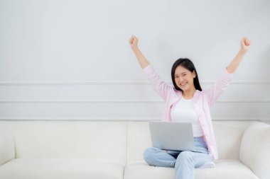 Happiness young asian woman sitting on sofa and working on laptop computer while successful and excitement at home, freelance female using notebook and achievement, business and lifestyles concept.