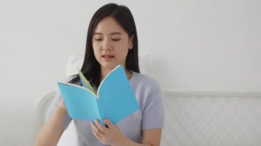 Genç Asyalı kadın rahatça kitap okuyor. Yatak odasındaki yatakta rahat rahat uyuyor. Kadın boş vakitlerinde kitap okuyor..