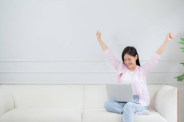Mutlu Asyalı genç bir kadın koltuğunda oturup dizüstü bilgisayarda çalışırken evde başarılı ve heyecanlı, not defteri ve başarıyı kullanan serbest bir kadın, iş ve yaşam tarzı konsepti.