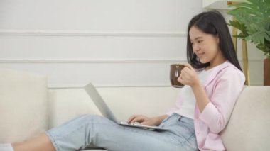 Young asian business woman work from home with laptop computer and drinking coffee on sofa in living room, freelance girl using notebook sitting on couch with comfort and relax, lifestyles concept.