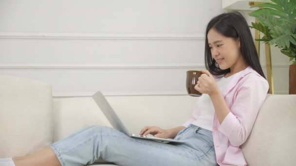 若いアジアのビジネス女性は 自宅からノートパソコンやリビングルームのソファでコーヒーを飲む 快適なソファに座ってノートブックを使用してフリーランスの女の子とリラックスし ライフスタイルの概念 — ストック動画