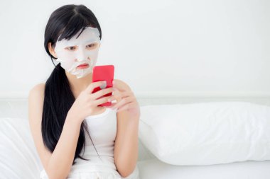 Yüzünde çarşaf maskesi olan güzel Asyalı bir kadın yatak odasında akıllı telefon izleyip cilt bakımı, cilt bakımı, sağlık ve sağlık için yüz maskesi takıyor..