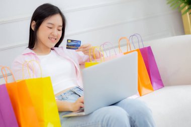 Kanepede oturan Asyalı genç bir kadın internet üzerinden dizüstü bilgisayar alışverişi yapıyor. İnternete kredi kartı alıyor, alışveriş, alım-satım ve ödeme ile ödeme yapan mutlu bir kız..