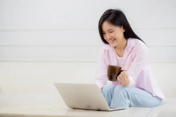 Young asian business woman work from home with laptop computer and drinking coffee on sofa in living room, freelance girl using notebook sitting on couch with comfort and relax, lifestyles concept.