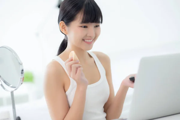Beauty of young asian woman with learning makeup with powder puff on cheek watching laptop computer with tutorial course online, female teaching and explain make up with cosmetic on streaming media.
