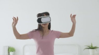 VR kulaklık takan genç Asyalı bir kadın evdeki yatak odasındaki yatakta heyecanla oyun oynuyor. Sanal gerçeklik ya da metaz yeniliklerini kullanarak 3 boyutlu, yaşam tarzları ve teknoloji kavramını kullanıyor..