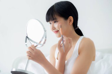 Güzel, genç Asyalı bir kadın. Yanak makyajına makyaj yapıyor. Yatakta kozmetik aynası, cilt bakımı ve yatak odasında kırışıklıklar olan güzel bir kız..