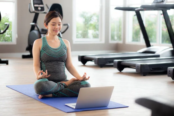 Young asian woman practicing yoga with watching laptop computer and meditating for relaxation in the fitness gym, female training yoga and exercise for bodycare and relax, rehab and health concept.