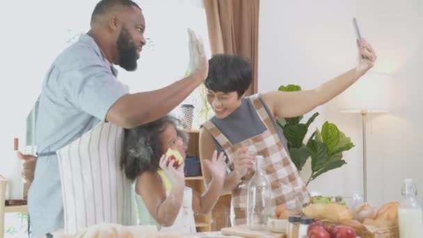 アフリカ系アメリカ人の家族の父親と母親が自宅のキッチンで娘と料理と一緒にスマートフォンで自撮り 父親と母親が一緒にスマートフォンで自撮り 家庭生活 ライフスタイルの概念 — ストック動画