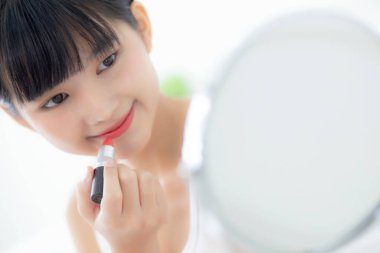 Güzel, genç Asyalı bir kadın dudaklarına ruj sürüyor, dudaklarına kırmızı ruj sürüyor, güzellik kızı makyaj malzemesi için aynaya bakıyor, zarif dudaklarıyla makyaj yapıyor, seksi bir kadın, cilt bakımı ve sağlık konseptiyle barışıyor..