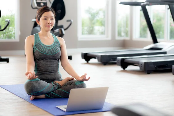 Young asian woman practicing yoga with watching laptop computer and meditating for relaxation in the fitness gym, female training yoga and exercise for bodycare and relax, rehab and health concept.